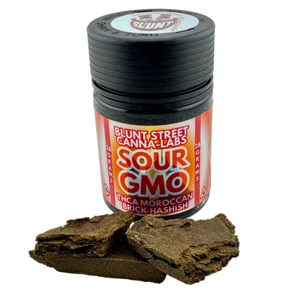 A black jar of Blunt Street Canna Labs Sour GMO THCA Moroccan Brick Hashish with a red and orange label, shown with several dark brown hashish bricks in front on a white background.