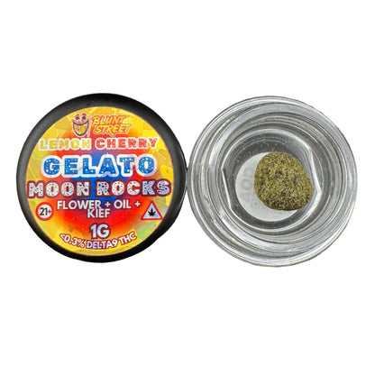 A small round black jar lid labeled 'Blingz Street Gelato Moon Rocks' sits beside an open clear glass jar containing a green flower oil concentrate puck on a dark gray textured surface.