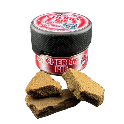 A small black jar labeled 'CHERRY PIE' with a red and white label sits on a dark gray surface, with two broken brown cake-like pieces placed in front of it.