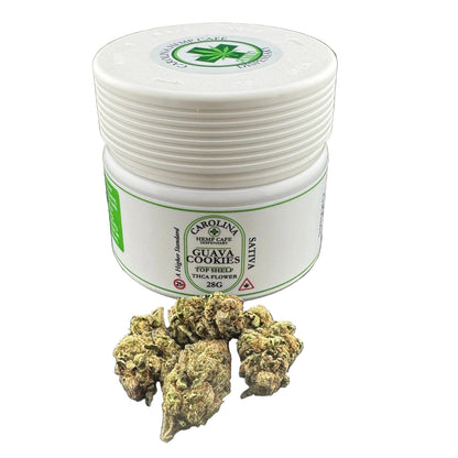 A white plastic jar labeled Carolina Hemp Co. Guava Cookies Top Shelf THCA Flower 28G, shown on a dark gray background with several green and brown dried flower buds placed in front of the jar.
