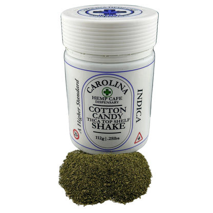 A white plastic jar of Carolina Hemp Cafe Dispensary Cotton Candy THCA Top Shelf Shake with a small pile of green hemp powder in front, set against a dark background.