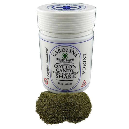 A white plastic jar of Carolina Hemp Cafe Dispensary Cotton Candy THCA Top Shelf Shake with a small pile of green hemp powder in front, set against a dark background.