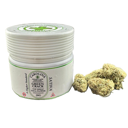 A white plastic jar of Carolina Hemp Cafe Green Crack Sativa THCA flower next to several dried green cannabis buds on a dark gray background.