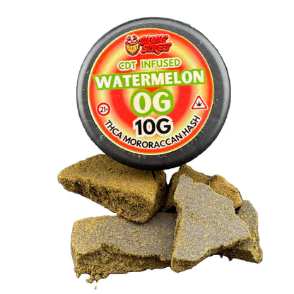 A round black container of GDT Infused Watermelon 0G 10G THCA Mororaccan Hash sits on a dark gray surface with several broken pieces of the brownish-green hash in front. The label is red, yellow, and green.