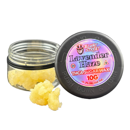 A small clear jar of sugar wax with its black lid placed beside it on a dark gray surface. The lid label reads 'Blunt Street Lavender Haze' and 'THCA SUGARWAX 10G'.