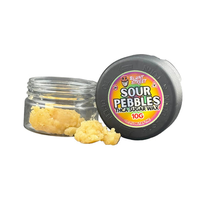 A small clear jar filled with pale yellow wax beads sits beside a black round tin lid labeled 'Sour Pebbles THCA Sugar Wax 10g' on a dark gray surface.